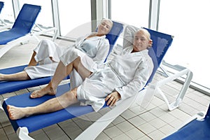 An elderly woman with a man in bathrobes on a sun lounger by the pool