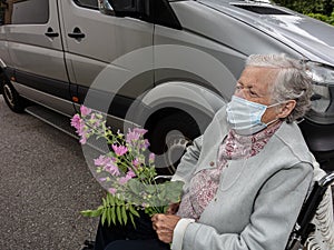 An elderly woman in a medical mask with a flower in her hands. Concept of a disabled person in a wheelchair transporting