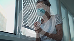 Elderly woman in a medical mask washing hands with alcohol gel or antibacterial soap sanitizer standing by the window in
