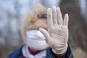 An elderly woman in a medical protective mask and rubber gloves gestures stop coronavirus