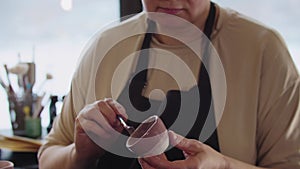 An elderly woman painting a little clay cup with dusty rose color and looks in the camera