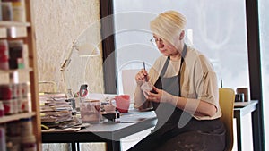 An elderly woman painting a little clay cup and looks in the camera