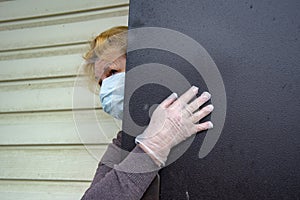 An elderly woman in a protective medical mask and rubber gloves looks out from behind a partially open door