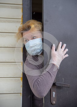 An elderly woman in a protective medical mask and rubber gloves looks out from behind a partially open door