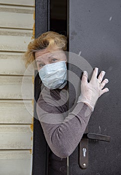 An elderly woman in a protective medical mask and rubber gloves looks out from behind a partially open door