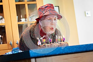 Elderly woman in a red floppy hat blowing out the lit candles on a birthday cake