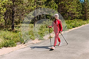 An elderly woman in red tracksuit walks with Nordic walking sticks in the summer