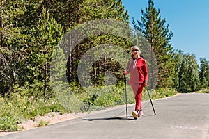 An elderly woman in red tracksuit walks with Nordic walking sticks in the summer