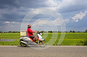 An elderly woman riding a motorcycle blurs through green rice fields