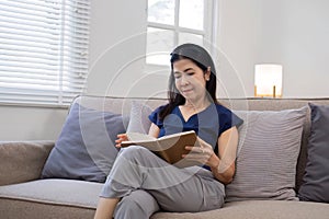 An elderly woman& x27;s leisure day sitting and reading a book, relaxing on the sofa in the living room.