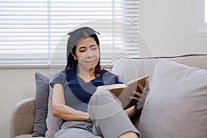 An elderly woman& x27;s leisure day sitting and reading a book, relaxing on the sofa in the living room.