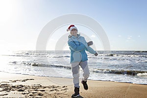 An elderly woman in a Santa hat on the seashore on a sunny winter day. Celebrating New Year and Christmas. Full height