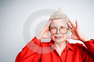 An elderly woman with short hair in glasses for vision holds her head with her hands from a headache, closing her eyes