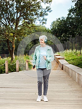 An elderly woman with a short haircut dressed in sportswear is jogging in the park while listening to music.