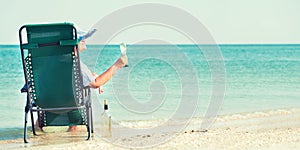 An elderly woman sits on the beach on a chaise longue and drinking wine.