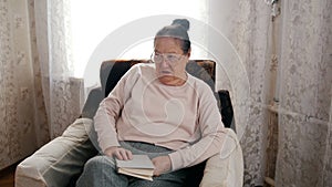 An elderly woman sitting in a chair and reading a book, looking with contempt.