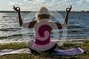 An elderly woman sitting in the lotus position and raising her hands up, on a mat in the park and looking at the lake