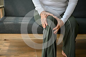 An elderly woman sitting on a sofa holding her knee, suffering from knee pain or joint ache