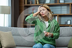 An elderly woman is sitting on the sofa at home and suffering from a headache, pressure, tension. holding his head