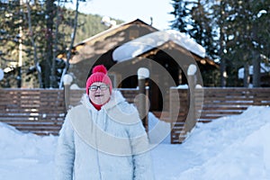 Elderly woman stands and smiles happily in front of a rustic wooden house among the snowdrifts in the forest