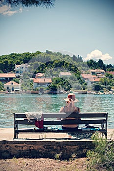 Elderly woman with straw hat sitting on bench by the sea and enjoying the view