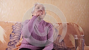 An elderly woman taking medication. Woman in the Age of sitting on the couch takes a pill, washed down with a glass of water.