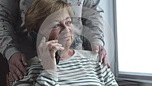 An elderly woman talking on the phone is upset and worried. Tears on eyes. Daughter supports her by hugging her