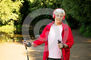 Elderly woman trains endurance while Nordic walking. Old female listening music in headphones.