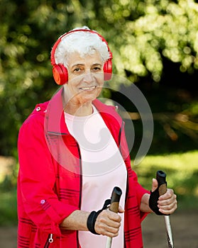 Elderly woman trains endurance while Nordic walking. Old female listening music in headphones.