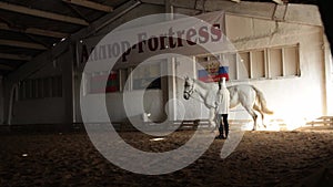 An elderly woman trains a horse on the arena. The inscription on the wall of Allur Fortress. A horse goes trot on the sand.