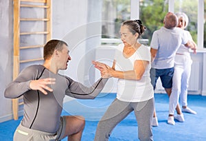 Elderly woman twists arm of attacking man with painful hold in gym. Self-defense lesson