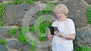 Elderly woman using tablet computer with earphones. Vintage wall of wild stone in the background.