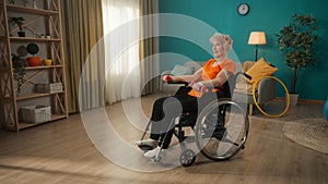 Elderly woman in a wheelchair exercising with dumbbells in the living room. An aged grayhaired woman trains arm muscles