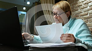 Elderly woman working at home with the documents using laptop.