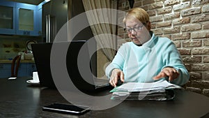 Elderly woman working at home with the documents using laptop.