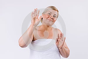 An elderly woman wrapped in a white towel with a smile poses and shows healthy and moisturized soft hands on a white