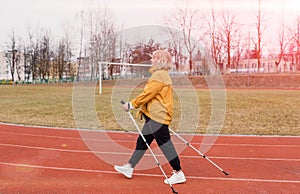 An elderly woman in a yellow sports jacket practices Nordic walking outdoors on the stadium`s rubber treadmill. A sunny sunset.