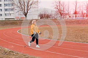 An elderly woman in a yellow sports jacket practices Nordic walking outdoors on the stadium`s rubber treadmill. A sunny sunset.