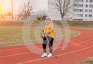 An elderly woman in a yellow sports jacket practices Nordic walking outdoors on the stadium`s rubber treadmill. A sunny sunset.