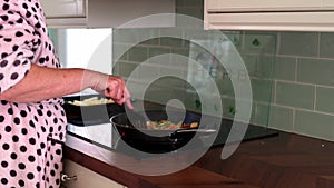 Elderly women in dressing gown stirning food on frying pan, cooking chicken chorizo