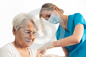Elderly worried lady with grey hair sitting and having an injection in her arm done by a female nurse in a blue medical