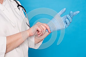 Eldery caucasian doctor lady in white coat and with stethoscope on blue background. Close up of putting on sterille gloves