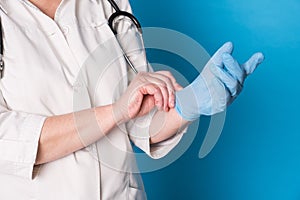 Eldery caucasian doctor lady in white coat and with stethoscope on blue background. Close up of putting on sterille gloves