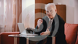 An eldery grandfather in glasses sitting by the laptop and looking in the camera