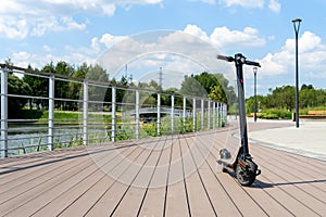 An electric black scooter stands on the bandwagon on the street. City park with wooden flooring along the promenade with railings.