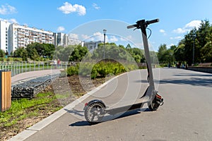 An electric black scooter stands on the bandwagon on the street. City park with wooden flooring along the promenade with railings.