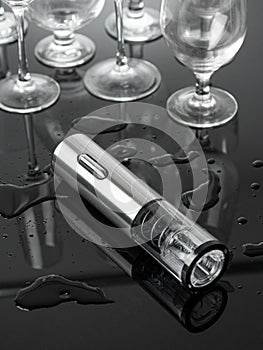 Electric corkscrew in gray metal. Lies on a mirrored surface. Empty glass goblets in the background