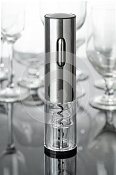 Electric corkscrew in gray metal. Stands on a mirrored surface. Empty glass goblets in the background