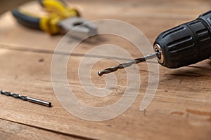 Electric drill bit and pliers lie against the wooden surface. The hand-held power tool is on top of the boards. Selective focus.