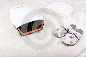 Electric heater and pair of warm slippers on white furry carpet. Winter background